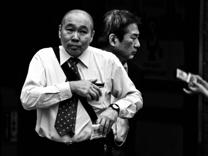 Two faces at Shimbashi station, one bald, one with closed eye including a pair of hands. Street Photography by Victor Borst