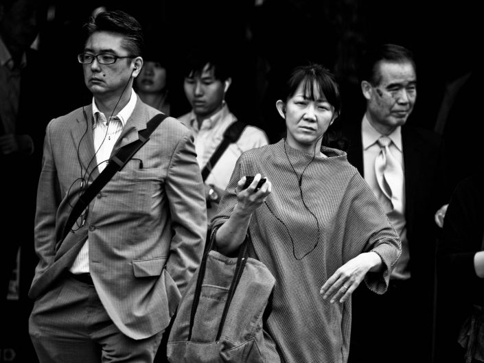 One Japanese woman and on man listening on headphones to music on their way to work. Street Photography by Victor Borst